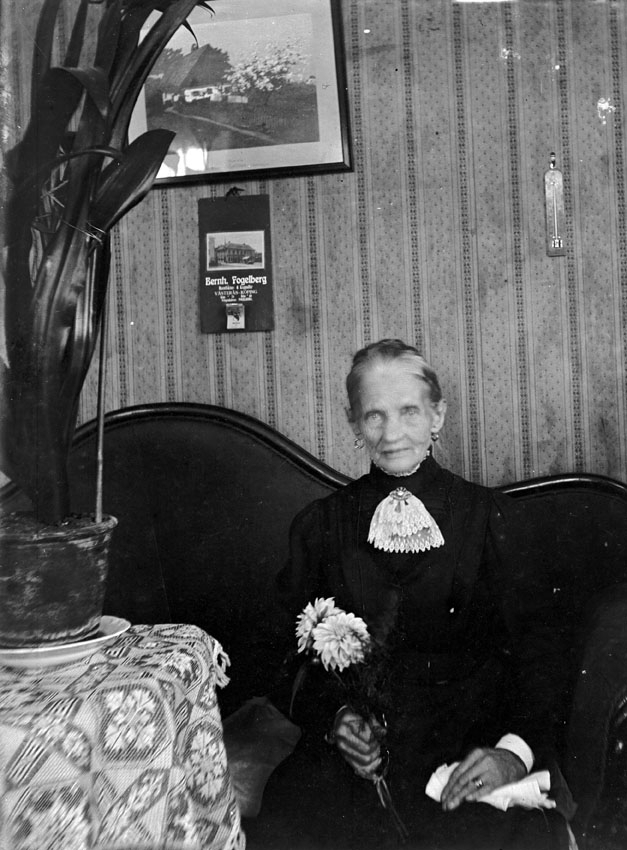 Äldre kvinna sitter i soffan med en blomma i handen.
