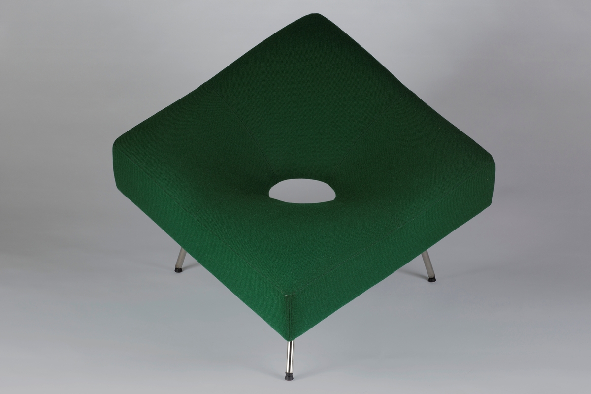 Lenestol formet som en kvadratisk pute med en sitegrop med et hull. Stolen har tre blankpolert ben i rustfritt stål og er trukket med et grønt ullstoff.  Den har en indre ramme i treverk polstret med formstøpt kaldskum og fiber.
