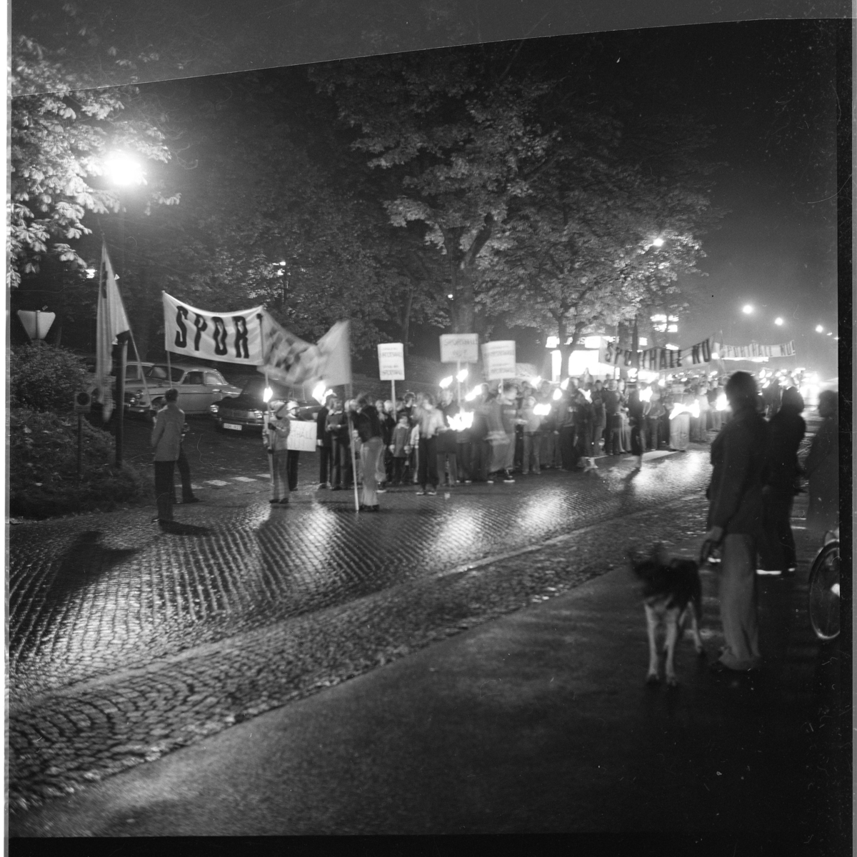 Ungdomar och vuxna med banderoll och plakat om "Sporthall" på Brahegatan i Gränna. Grännabor demonstrerar för att få en sporthall till Gränna. Det är oktober, mörkt och blött på vägen.