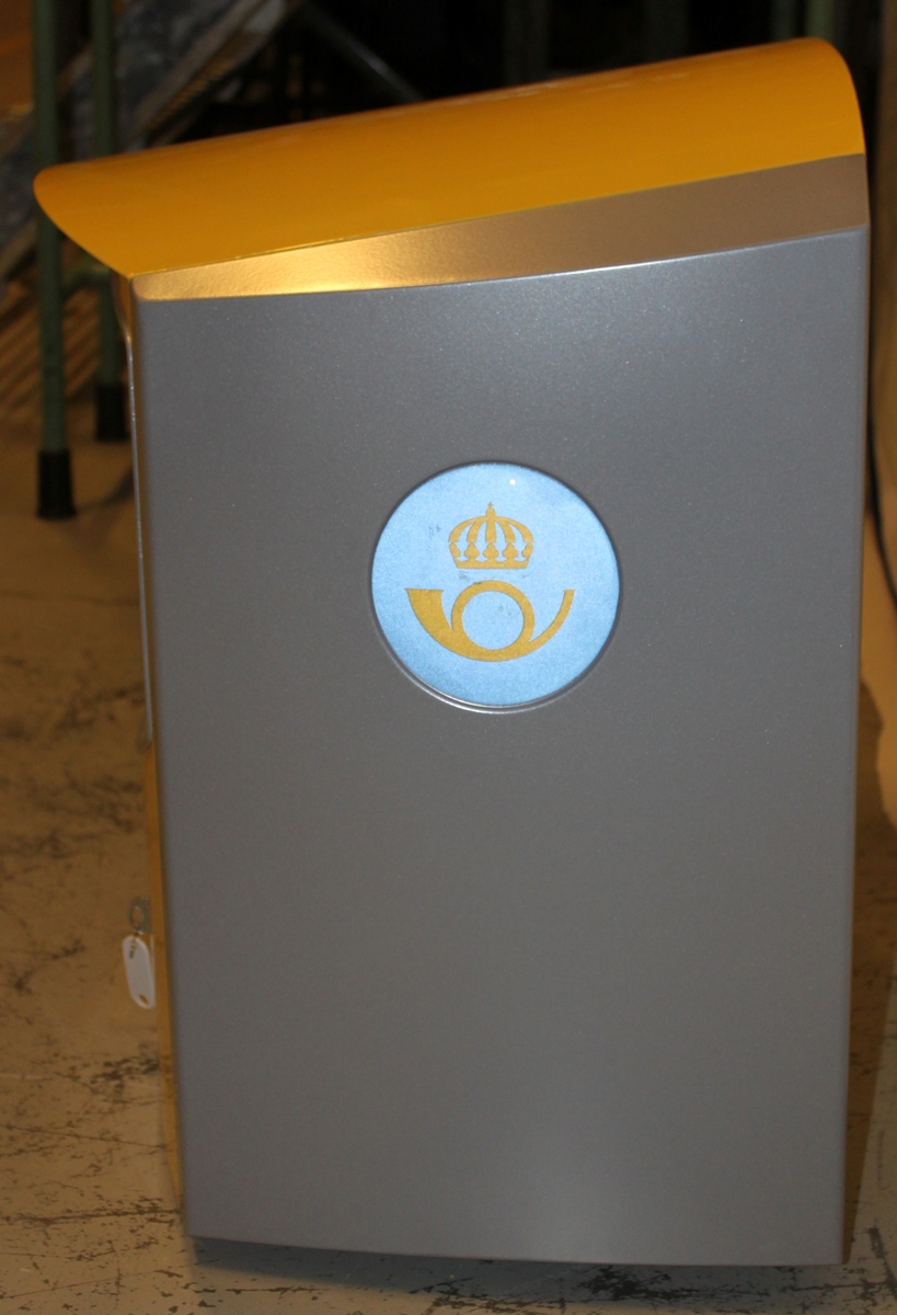 Brevlåda av den typ som infördes 2001-2002, gul med silvergrå sidor. På sidorna finns även Postens nya symbol ett gult krönt posthorn på en ljusblå cirkel. Brevlådan töms från fronten. Nyckel finns.