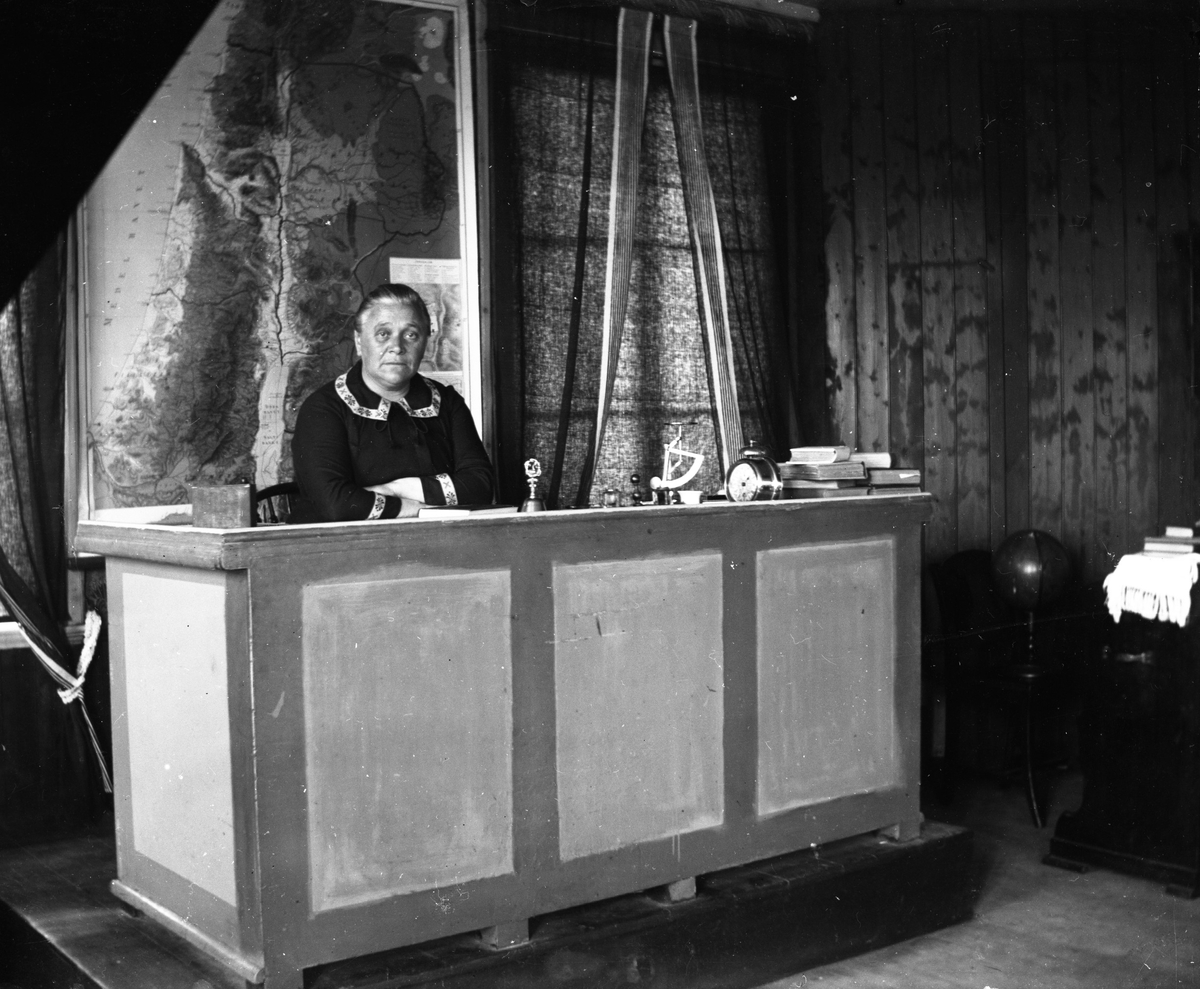 Folkskollärarinnan Constance Engström i Sällstorp sitter i katedern. Hon innehade tjänsten åren 1891-1921. Bakom henne hänger en stor karta och på katedern finns, förutom böcker, en ringklocka, en våg och en stor väckarklocka. Till höger står tramporgeln och en jordglob. Constance var moster till fotografen.