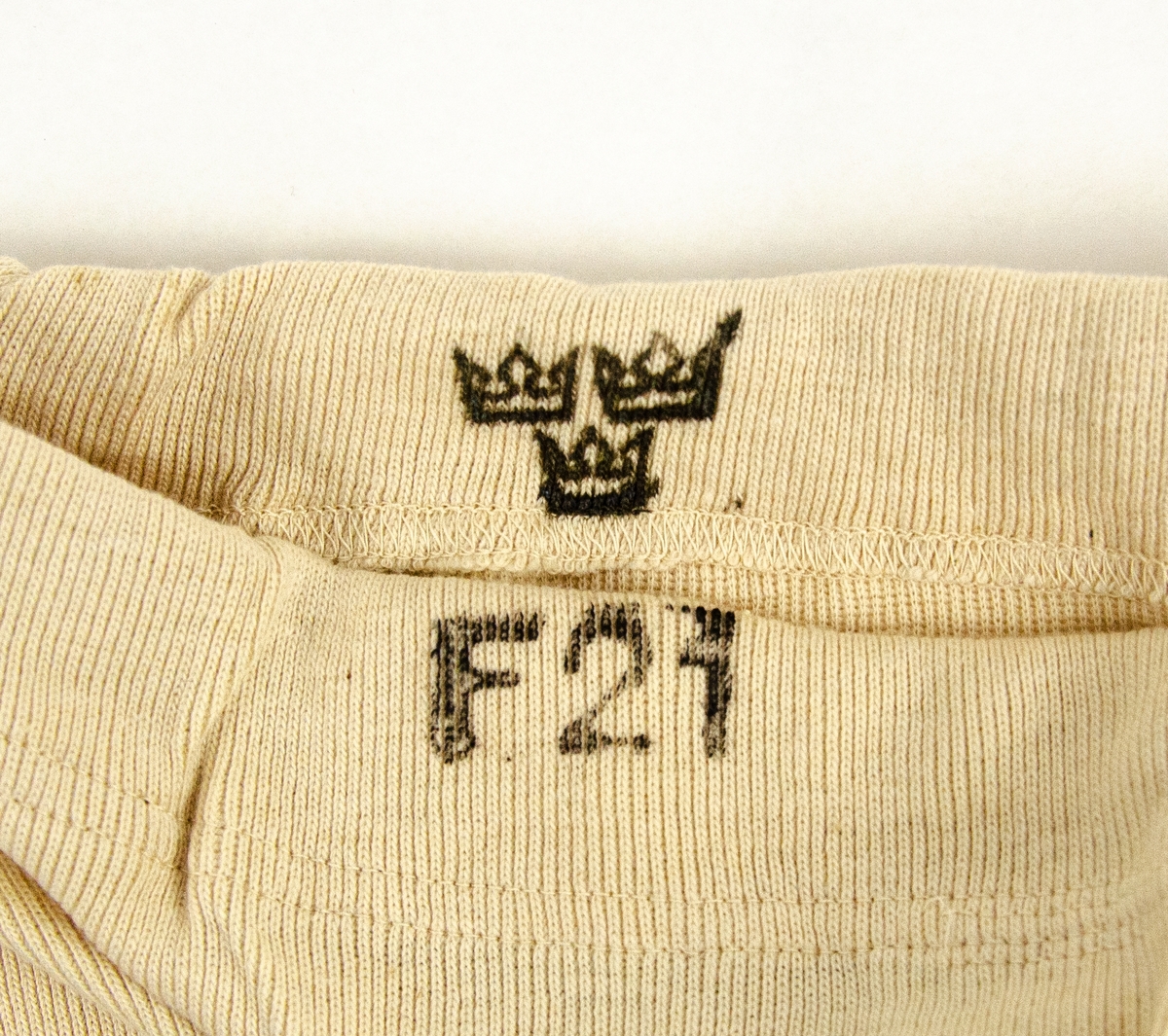 Underställsbyxa utförd i oblekt bomullstrikå. På insidan av byxlinningen finns maskingjord etikett "storl 5.", samt stämplat motiv i svart bläck iform av tre kronor. På byxlinningens utsida finns i svart bläck stämplat "F21".