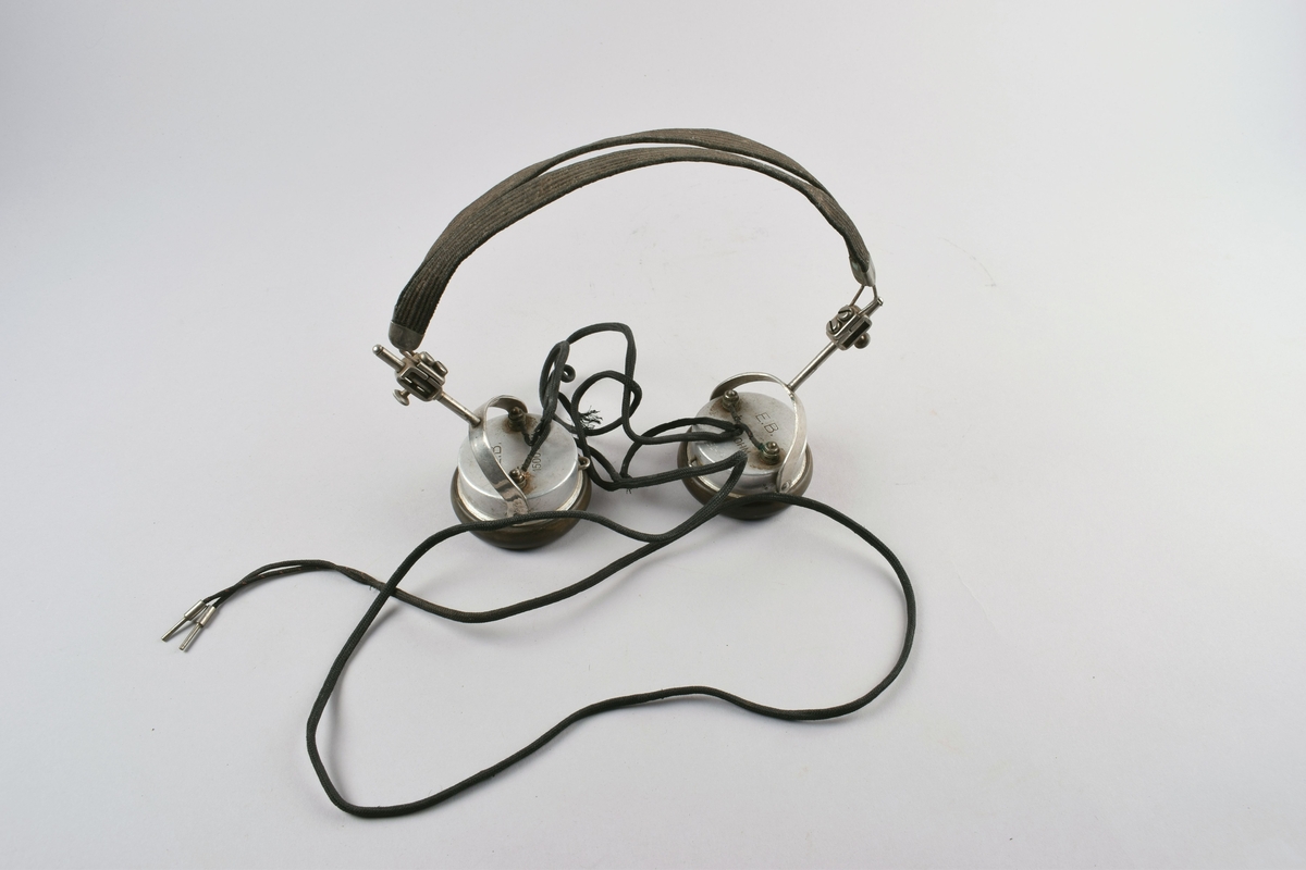 Høretelefoner forbundet med dobbel hodebøyle i metall trukket med tekstil. Ledning som splittes i to plugger nederst. Har vært brukt sammen med krystallapparat.