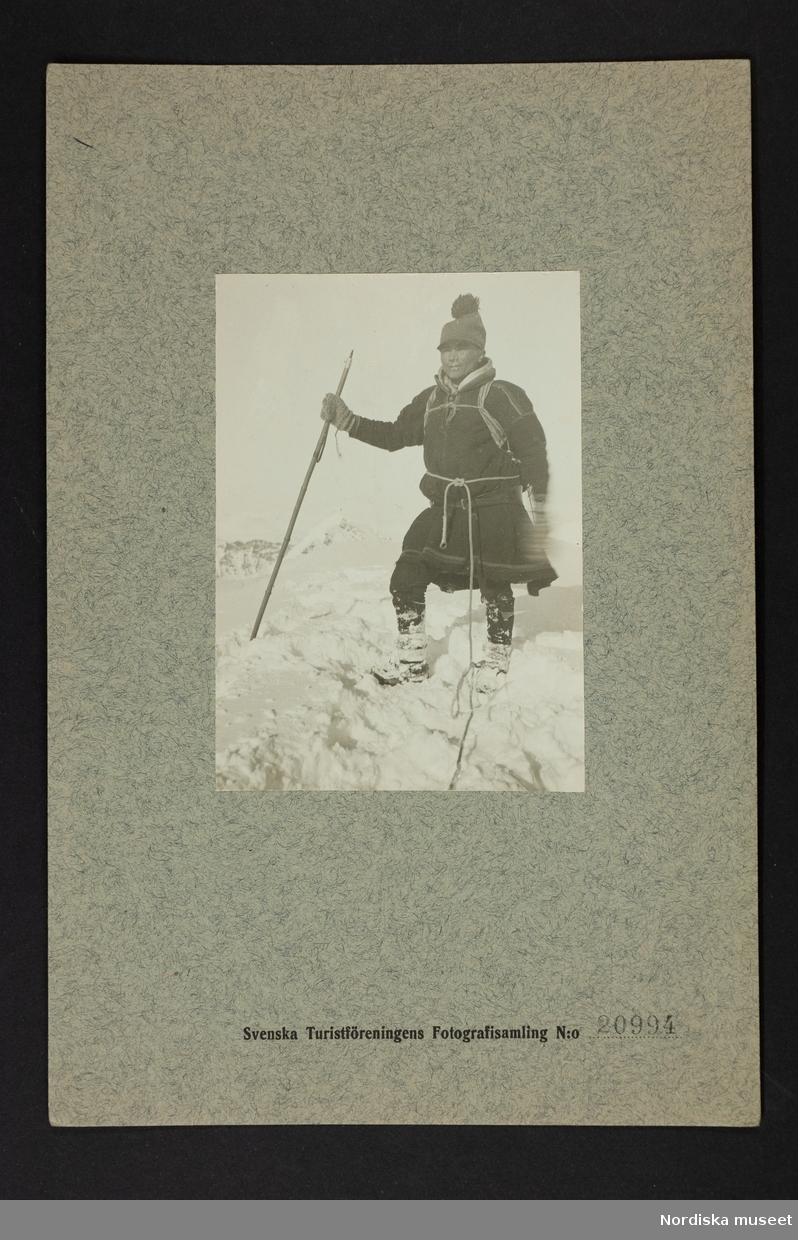 Porträtt av samisk man stående i snö med stav i handen. Påskrift "Kebnekajse Nia på toppen 29 juli 1906".