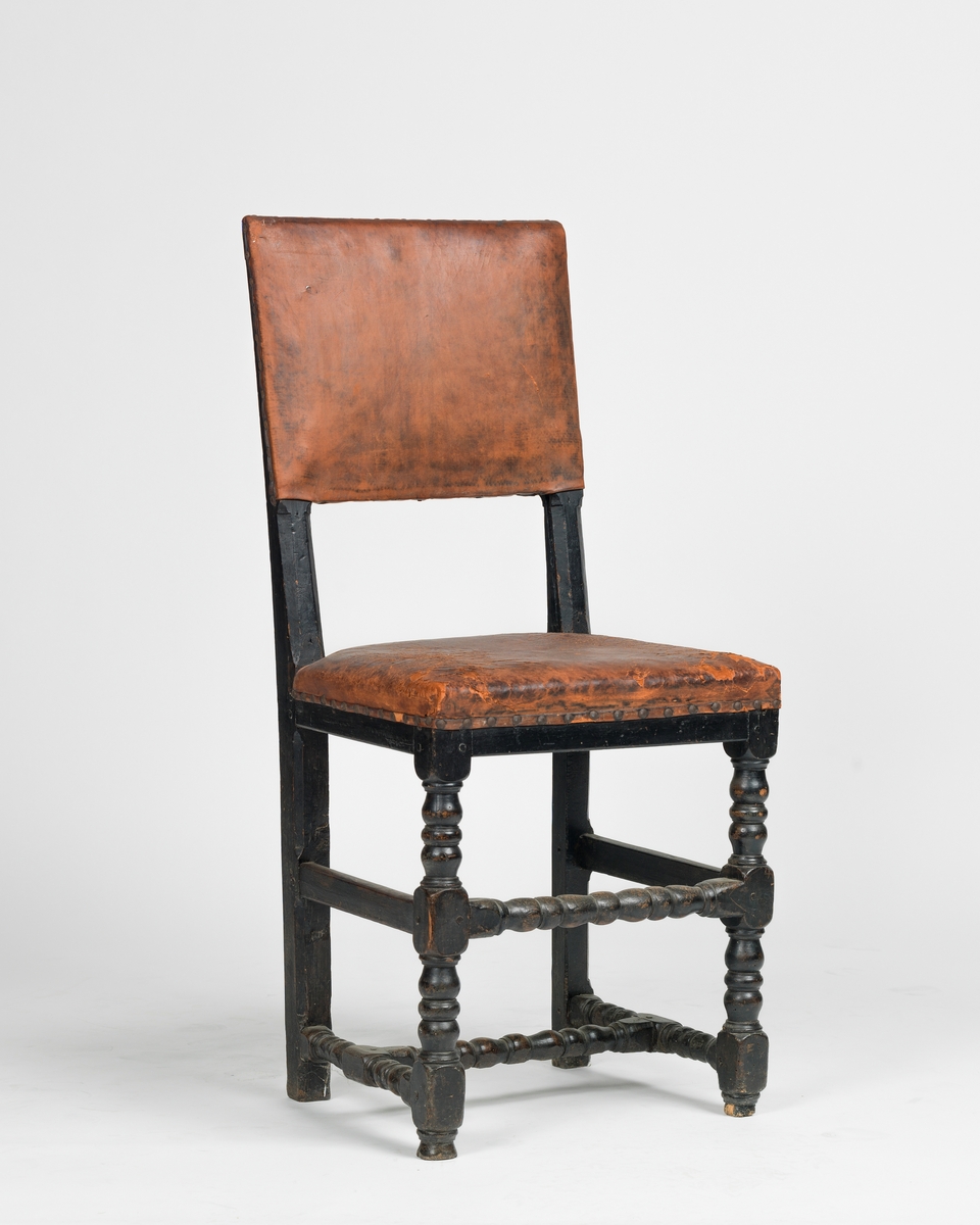 A-B: 2 stk. stoler i barokk stil med skinntrekk. Dreide ledd på de to fremste bena, for- og bakben sammenbundet med sidesprosser, rygg og sete stoppet og skinntrukket, malt. Rett sarg, rygg og sete, rektangulært ryggfelt.
