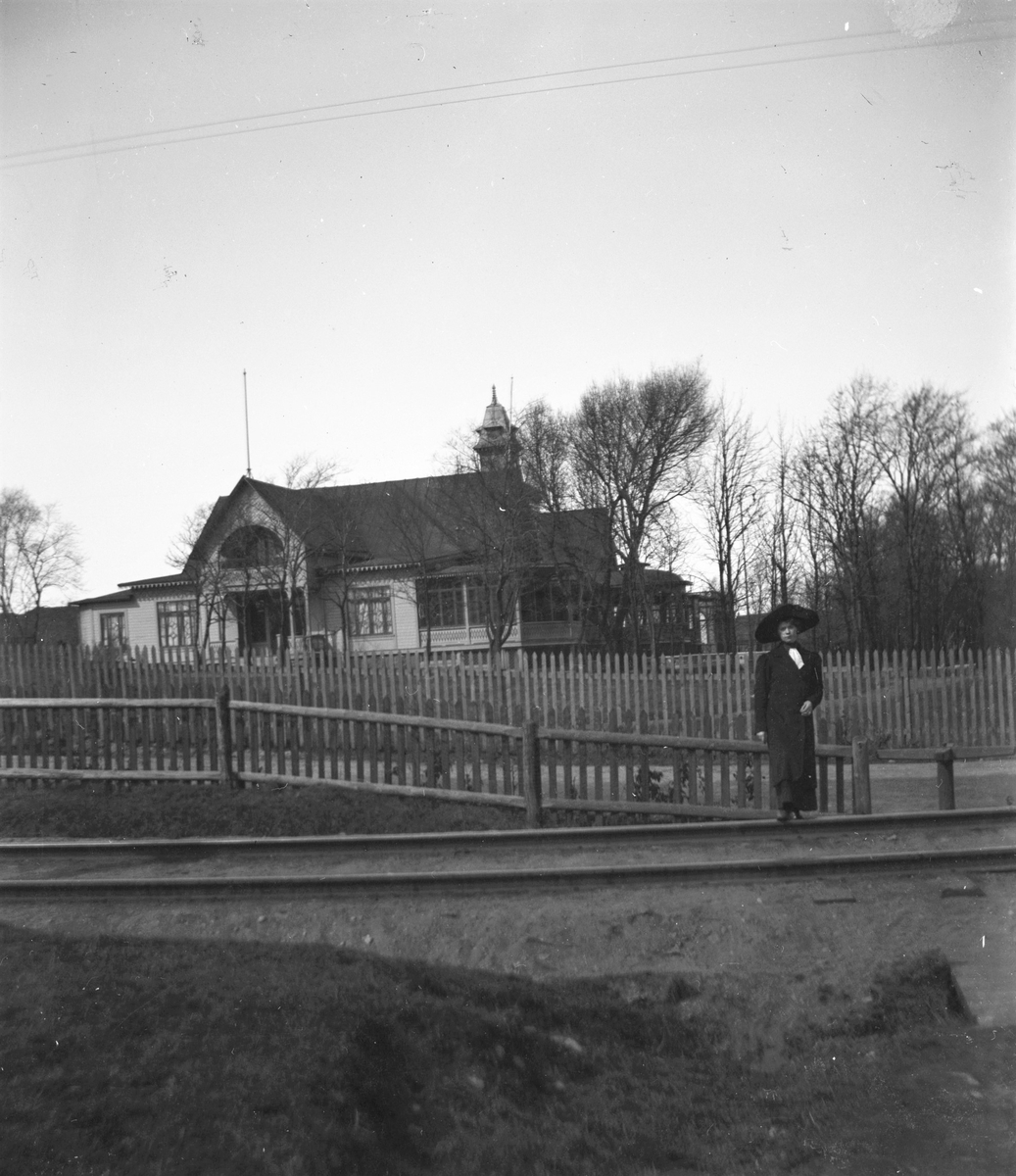 En ung kvinna står med ena foten på rälsen som ledde tågen söderut från Varberg före 1920. I bakgrunden ses Societetsrestaurangens gavel och spjälstaketen längs grusvägen däremellan.