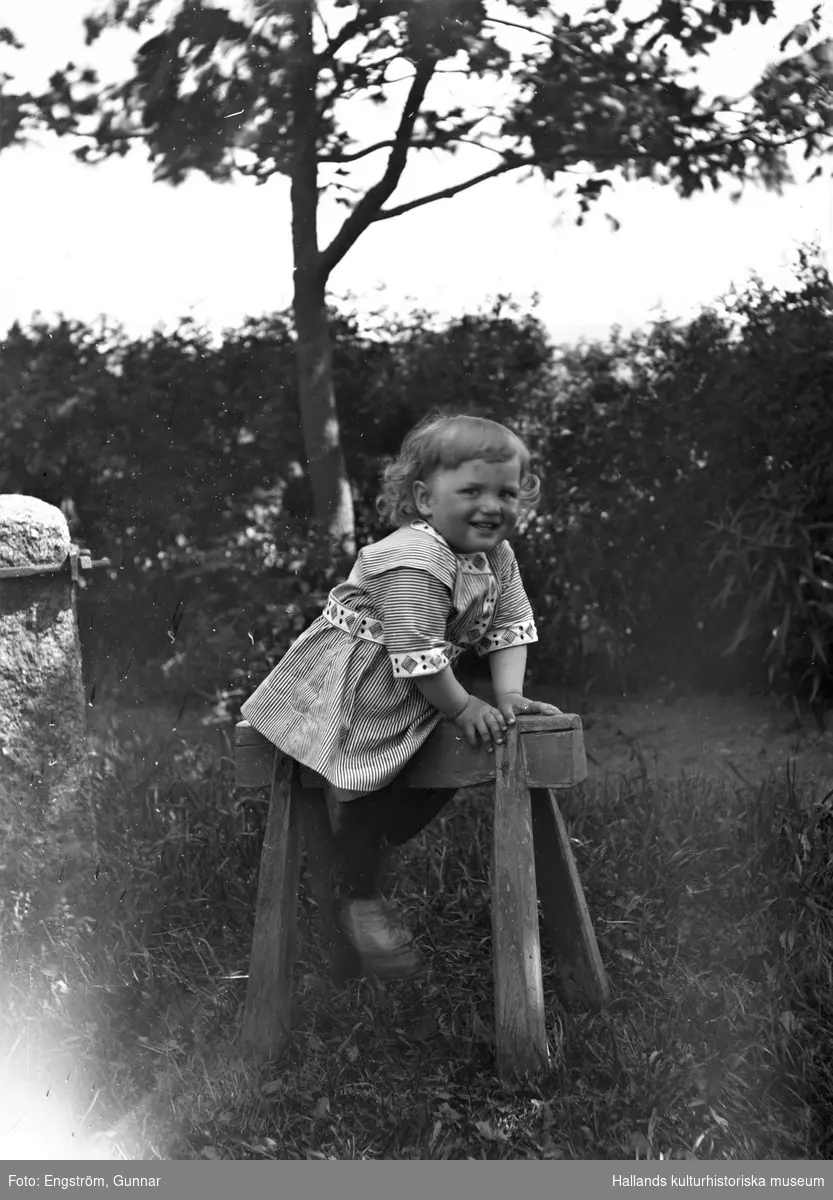En liten pojke i randig kolt sitter på en träbock i en trädgård. (Se även bildnr GEA136)