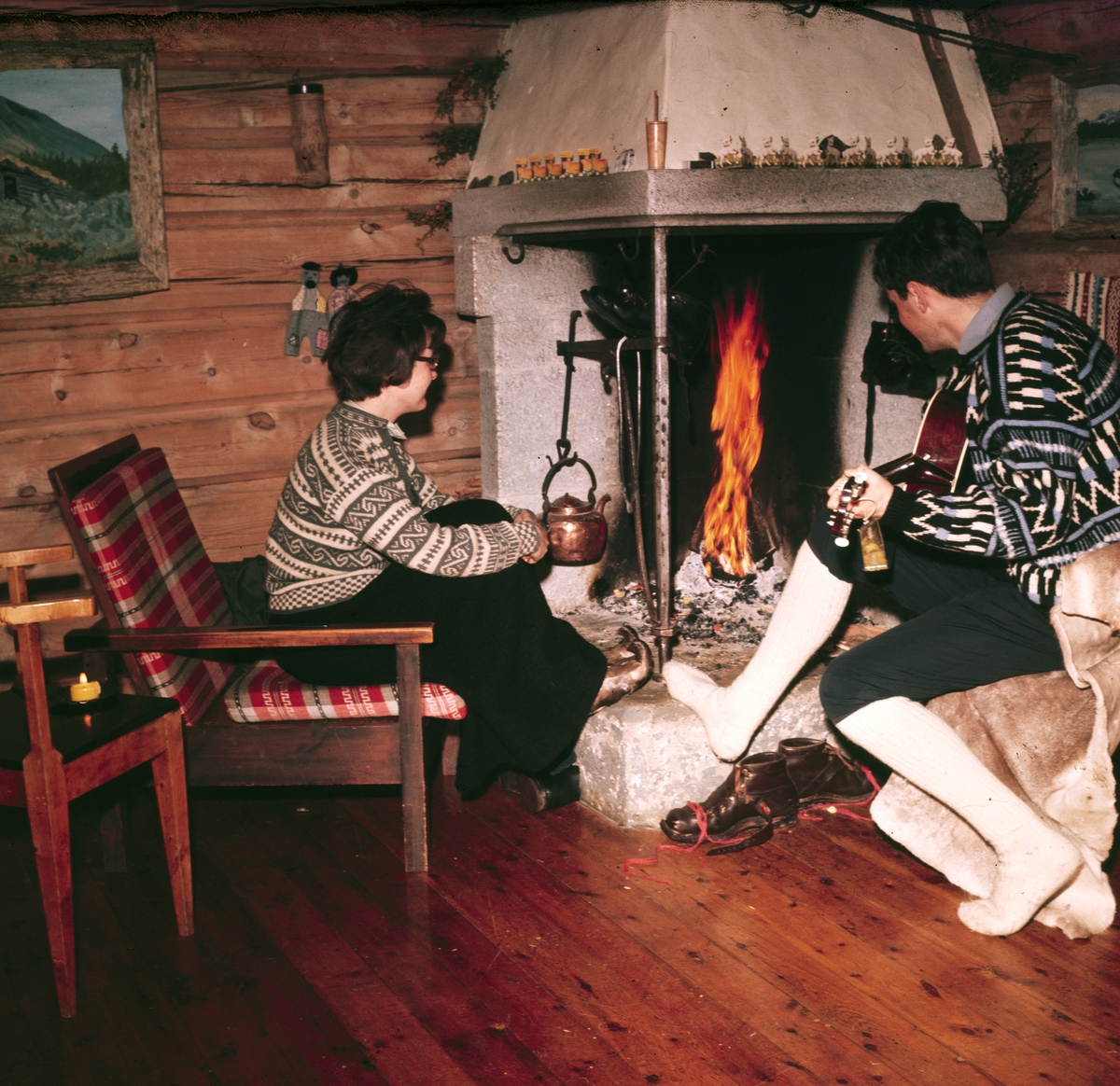Foto av et par som sitter foran peisen i en hytte. Han spiller gitar. De er kledd i strikkekofter begge to. Hytta er en tradisjonell laftet tømmerhytte med enkelt møblement.