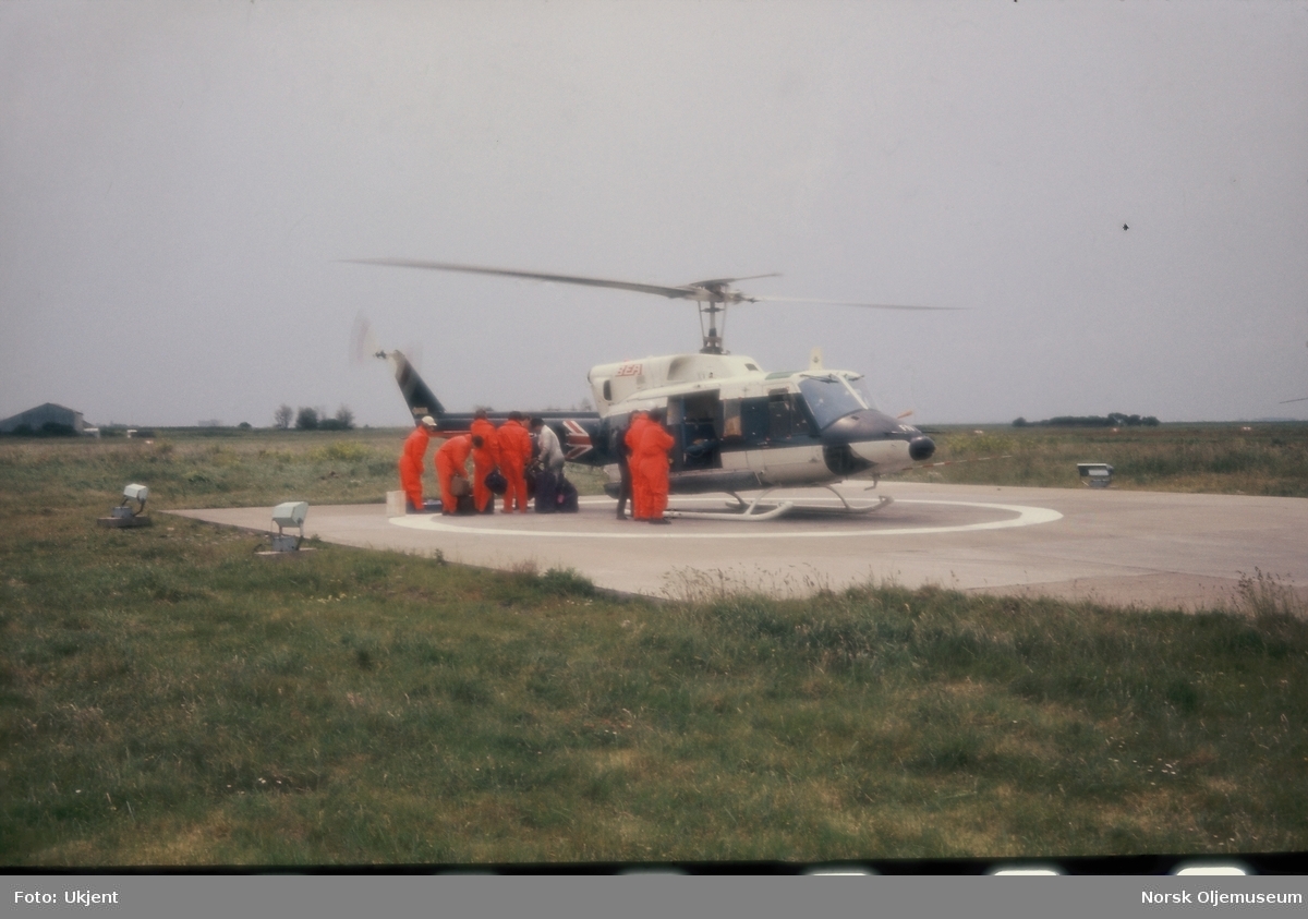 Arbeidere gjør seg klare for transport med et britisk BEA helikopter. Alle er kledd i orange arbeidsdresser.