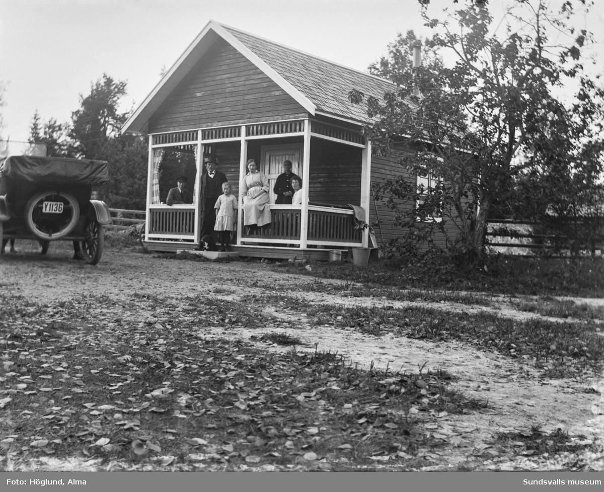 Fem vuxna personer, ett barn och en hund på verandan till en mindre stuga. Intill står Gottfrid Höglunds T-Ford 1922.