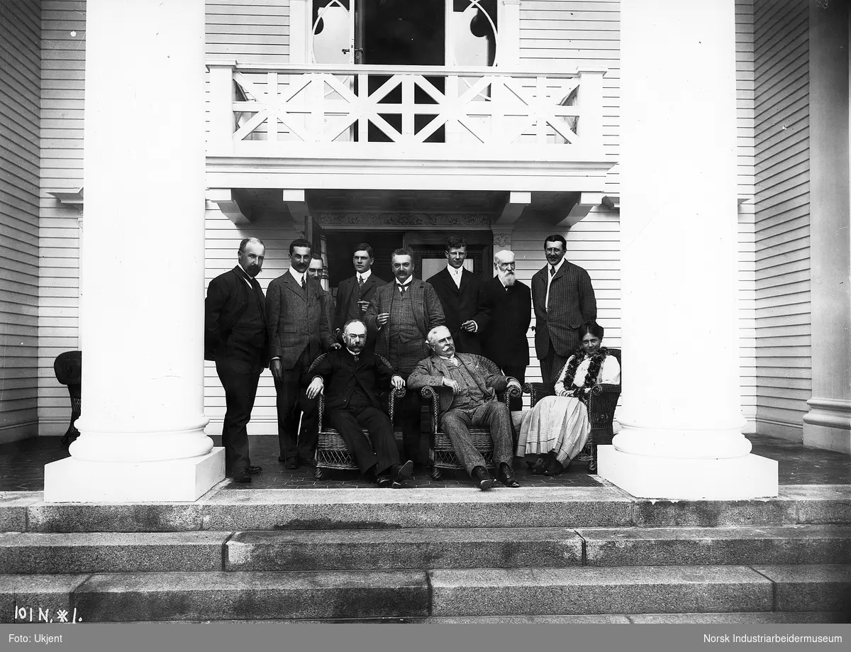 Sam Eyde med gjester på verandaen utenfor Notodden Admini. To menn og en kvinne sittende i kurvstoler foran og 8 menn stående bak, Sam Eyde i midten.