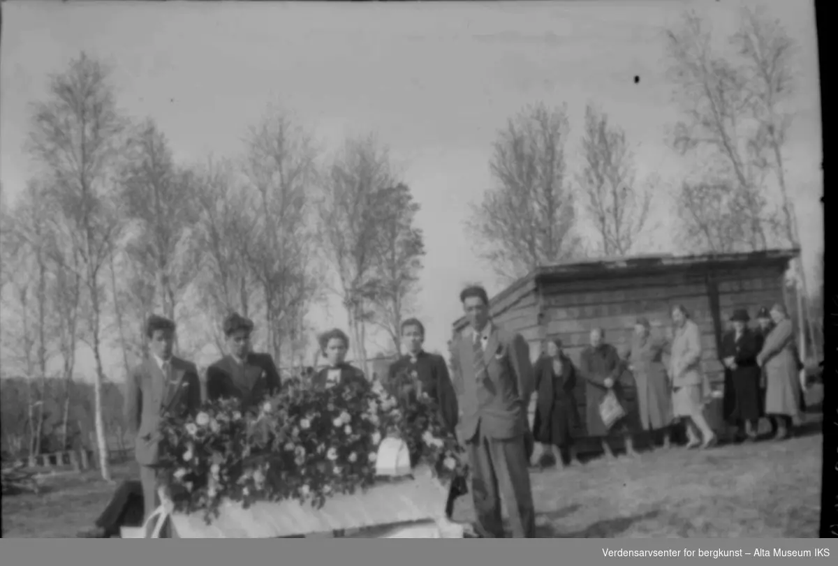 Begravelse med kiste dekket av blomster og familie samlet rundt.