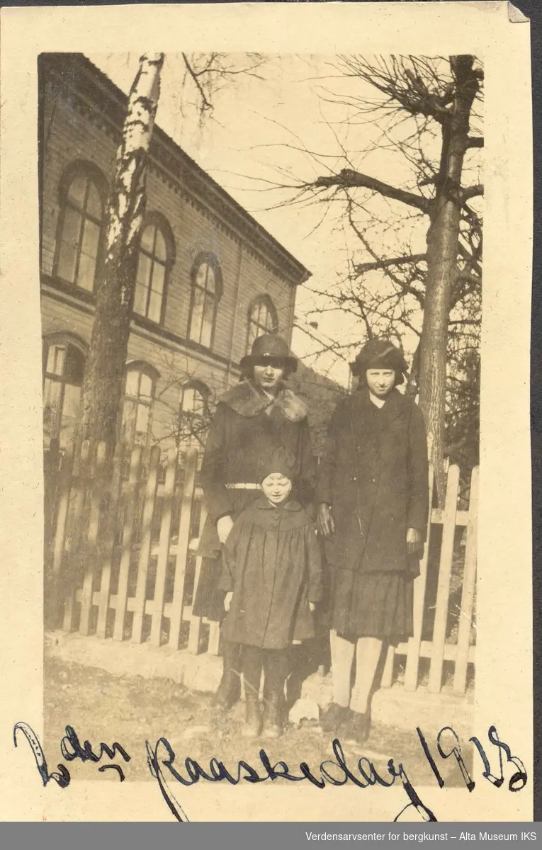 En voksen kvinne med en liten jente og en litt eldre jente foran et mursteinshus, trær og gjerde i bakgrunn. På bildet står det skrevet 
"2. Paaskedag 1923"
