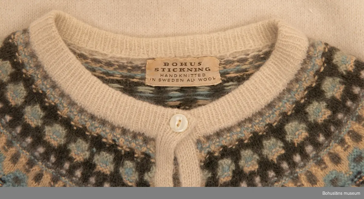 Stickad kofta med lång ärm, stickad enligt "gamla rundstingningen". Jumpern är slätstickad i de enfärgade partierna med resårstickad halsringning, muddar och nederkant. Oket är mönsterstickat med räta och aviga maskor i nyanser av blått, grått och grönt, mot en ljus beige botten. Knäppning fram med tio syntetiska knappar. Originaletikett: BOHUS STICKNING HANDKNITTED ALL WOOL