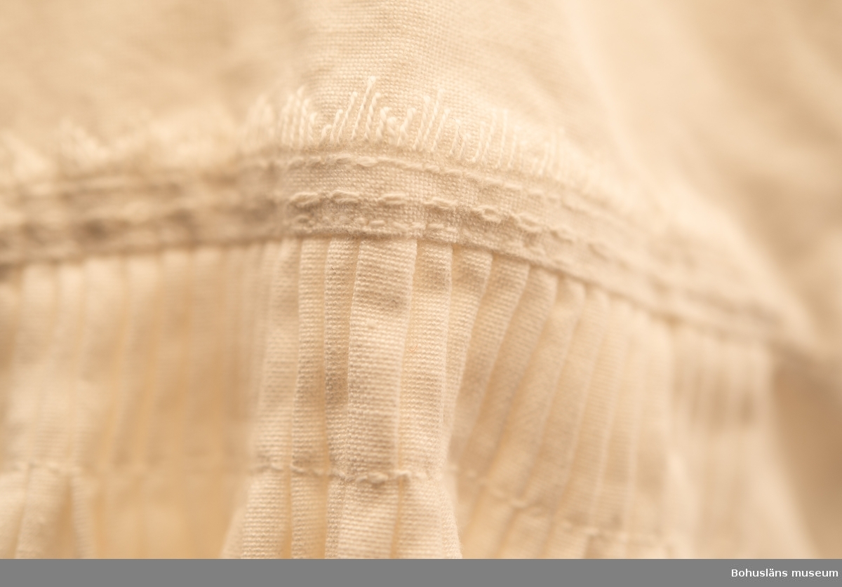 Del av bohusdräkt.
Knälång överdel, upptill av vit bomull och nederdel av oblekt linne.  Stort halshål utan knäppning. Överdelen har nedliggande ca. 10 cm bred rundad krage med lagda veck runtom, nederkanten dekorerad med hålsömsbroderi och smal knypplad spetskant. 
Isydda raka ärmar med ärmkil, axelparti med lagda veck och nertill lagda veck mot 6 cm breda manschetter, avslutade med små vikta uddkanter. Vitbroderier i olika mönster över axelparti och manschett. Ärmsluten med bandknytning.
Märkt I L-O 1981 i vitbroderi.
Mycket välsydd.