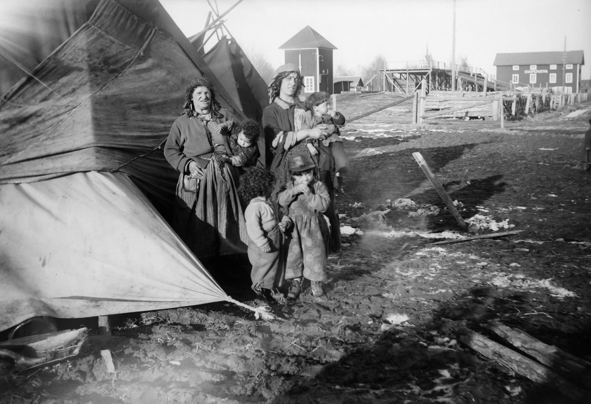 En familj poserar för kameran framför sitt tält. I bakgrunden skymtar Mockfjärds kyrka. Bilden är enl. uppgift tagen i mars 1929. Boendestandarden bland svenska romer var under 1900-talets första hälft mycket varierande.