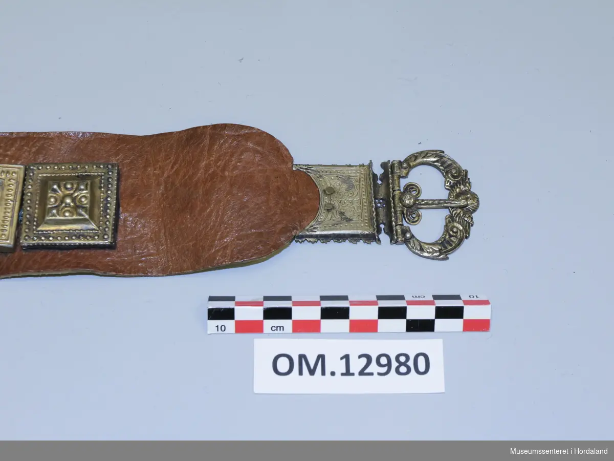 Stølabelte av brunt ler og forgylte sølvstølar og spenne i eine enden. På andre enden er ei hempe til å hekta spennenåla i. Stempel på baksida av spennet, 16 og 18 med romartal, L.K og L.N. Engelmotiv ytst. Stølane er ikkje stempla på framsida. Snor på baksida.