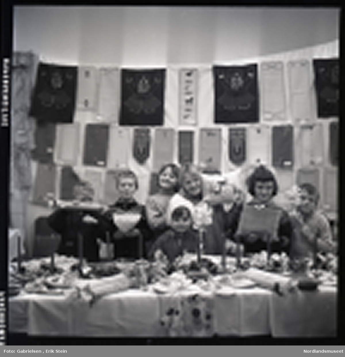 Fotografi av en gruppe med barn som selger puter med mønster og julelys og kurver og kjevler og juleduker og noen tøfler og andre ting på et langt bord i et rom med et vindu med noen gardiner et ukjent sted