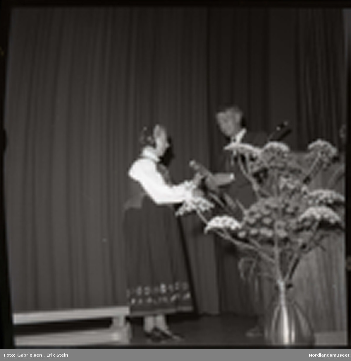 Fotografi av en eldre kvinne kledd i en bunad og en bunadshette som står på en scene i en sal og får utdelt en pris ev en mann kledd i dress med slips som står vedsiden av en talestol med en mikrofon og en stor blomsterkrukke 
som det vokser mange blomster i og en benk som står på scenen og man ser en rekke av blomstermønster på
bunaden til kvinnen i skjørtet på bunaden og bunadshetten