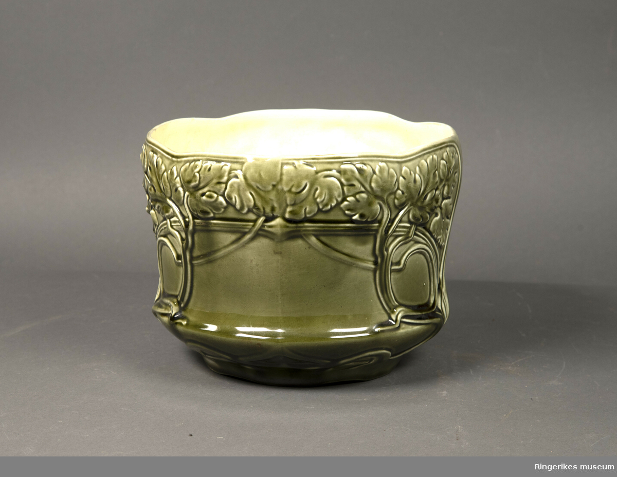 Stor potteskjuler i keramikk. Dekorert med ranker med blad som minner om vindrueblader. Glasert. Olivengrønn på utsiden, svært lys gul på innsiden.