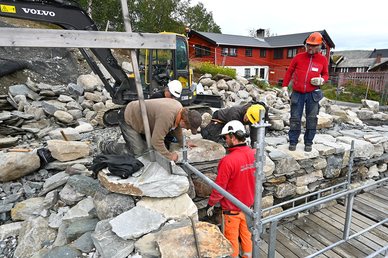 Bildet viser arbeidere som jobber med å bygge opp en større mur. I bakgrunnen ser man et rødt hus og grå himmel. (Foto/Photo)