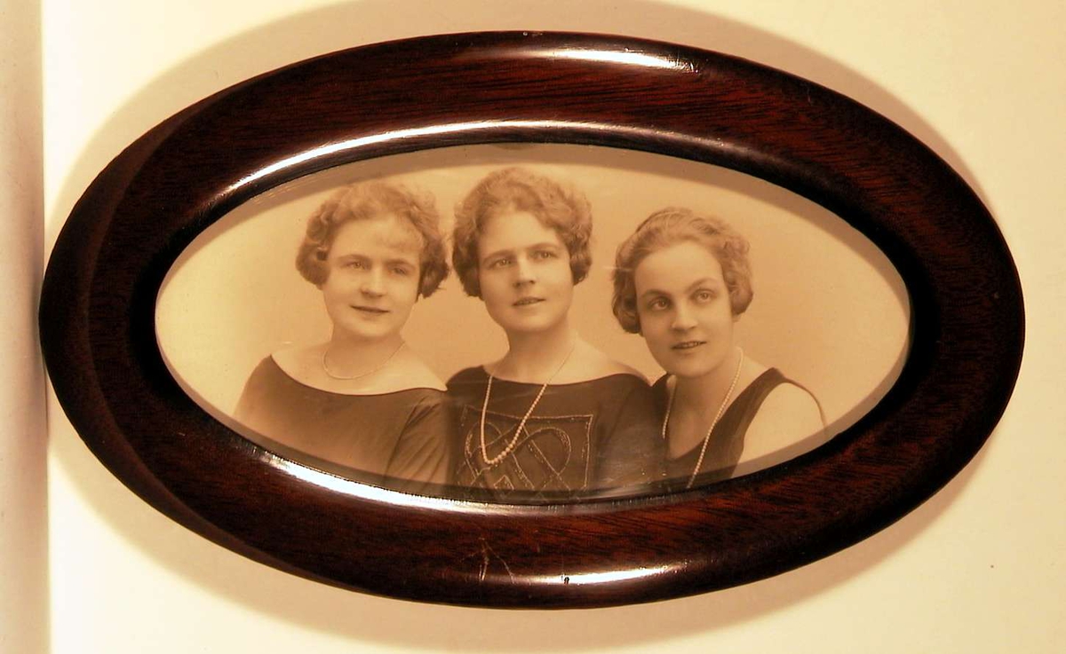 Atelierfotografi; familiebilde av tre unge damer i festkjoler, perlekjeder rundt halsen og fine korthårsfrisyrer lagt i bølger inn mot ansiktene.
Brystbilde i oval bredde-ramme.