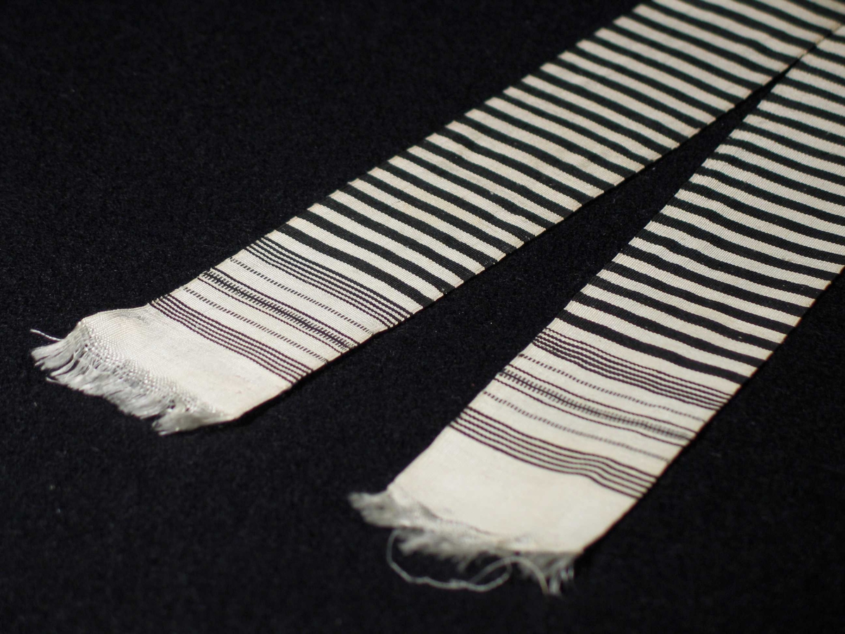 Tverrstripete silkebånd i svart og hvitt. Hvite frynser i begge ender dannes av renningen. Innslagsutglidning ved frynsene. Vevbredden er 4,5 cm.