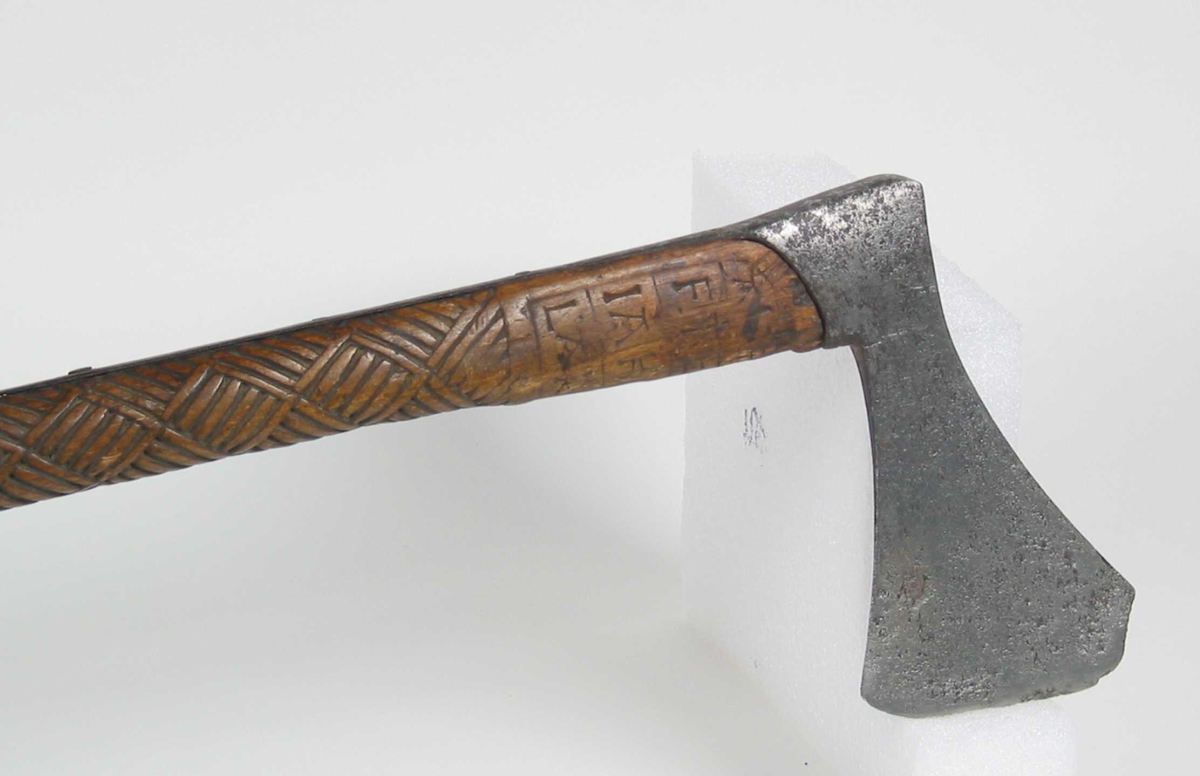 Asymmetrisk økseblad, sammensveiset med overliggende jernskinne.
Fremre øksebladspiss brukket.
Rikt dekorert skaft med flettet rutemotiv. Innskripsjon på øvre del av skaftet (se Påført tekst)
