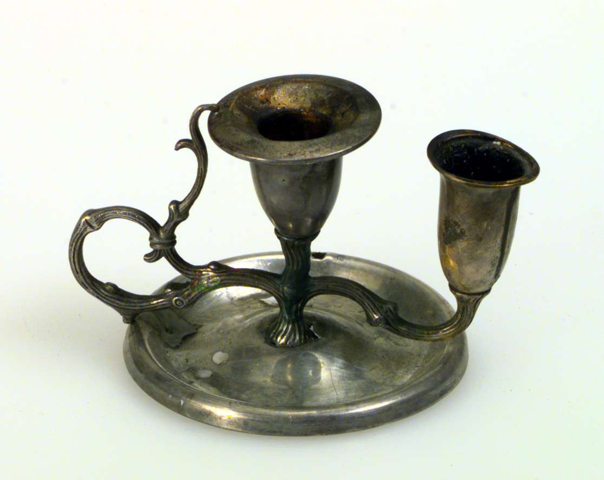 Lysestake med to piper og håndtak. Staken har rund fot. Foten er antagelig av sink, skaft med piper er av sølv. 