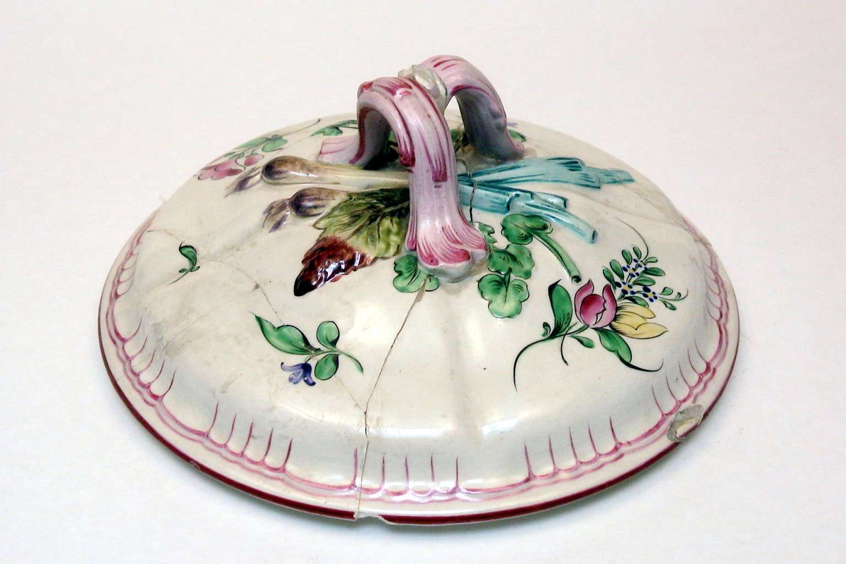 Rundt lokk i kremfarget keramikk med polykrom blomsterdekor. Lokket er defekt.
Fatet eller terrinen som lokket har hørt til, mangler.