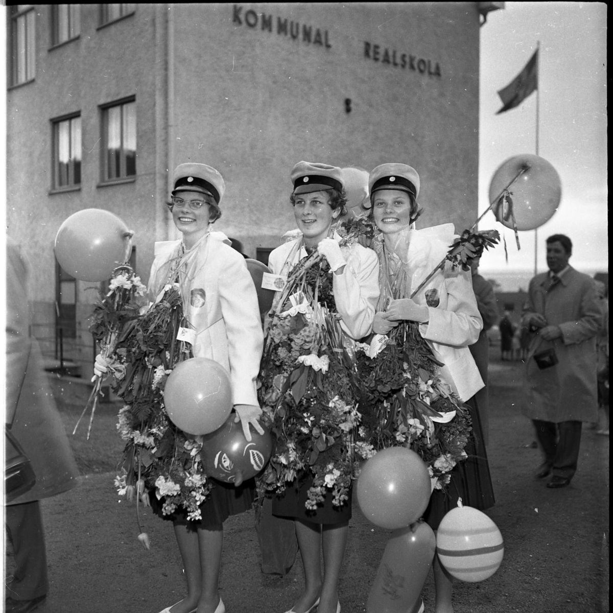 Realexamen 1961. Tre kvinnliga studenter står uppställda för fotografering. De bär realskolemössor och de har buketter och ballonger runt halsarna. I bakgrunden syns skolbyggnaden, på fasaden står det kommunal realskola.