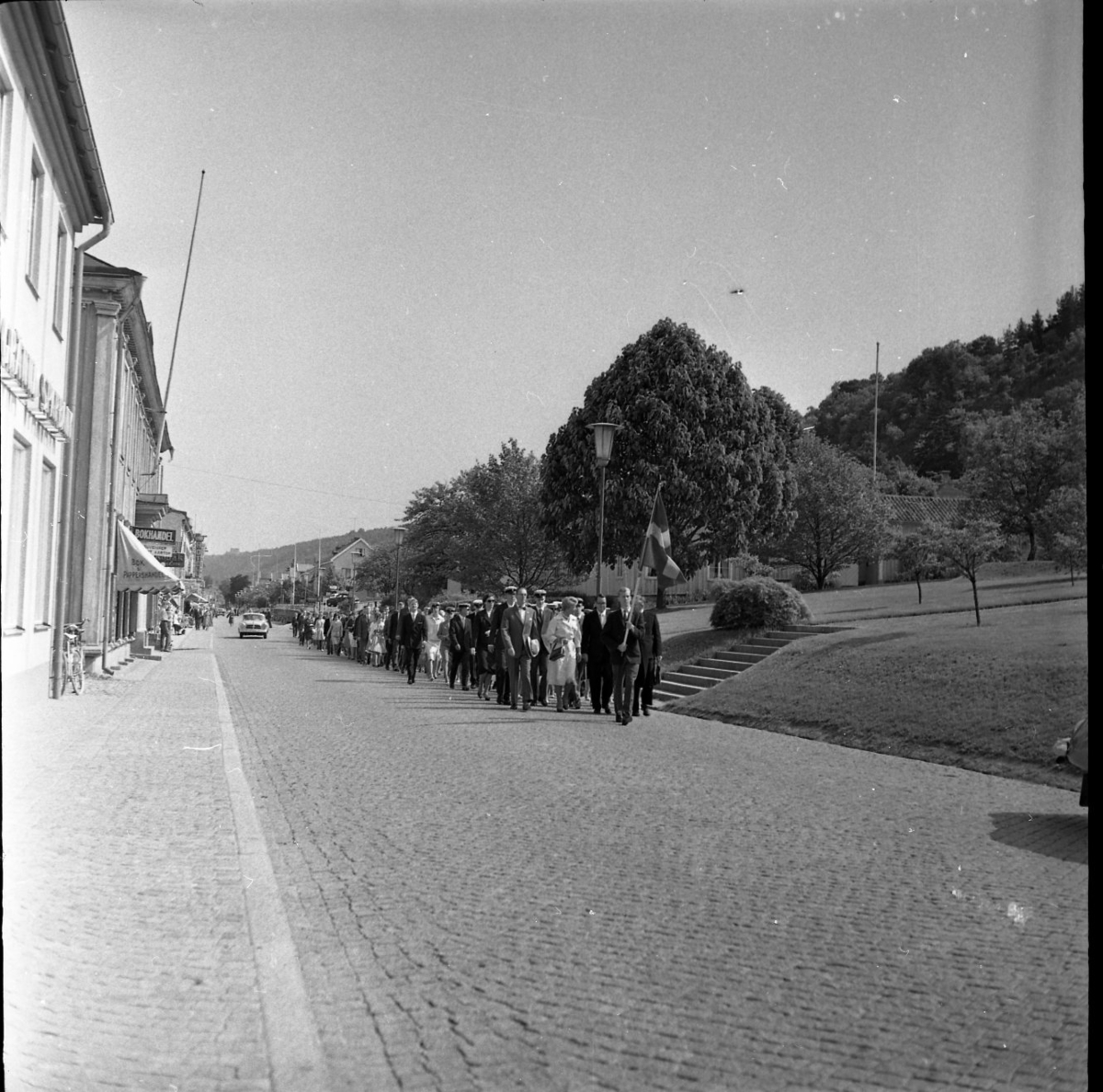 Studenter iklädda realskolemössor promenerar på ett led längst Brahegatan vid Braheparken. Längst fram går en fanbärare med svenska flaggan i händerna. I bakgrunden skymtar Franckska gården.