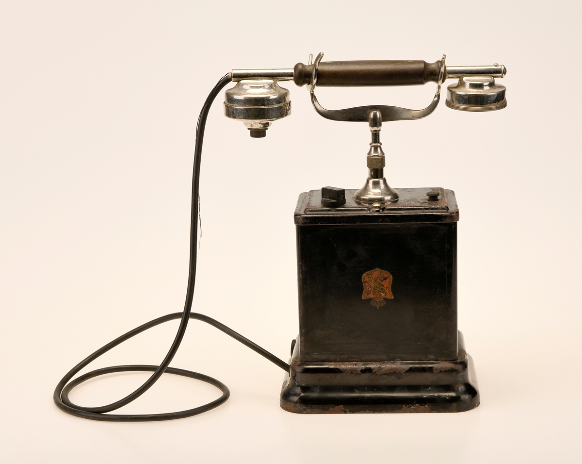 En svart bordtelefon av Elektrisk Bureau sin 1922 modell. "Den norske løve" pryder forsiden og baksiden av aparatkassen, disse er slitt og i tillegg oppned. Forøvrig har telefonen en y-gaffel. Ledningen som går fra aparatkassen til telefonrøret er av rund plast, noe som tyder på at den har blitt byttet ut på et tidspunkt. Sveiven mangler, deler av telefonrøret mangler, og ledningen med støpsel er fjernet.