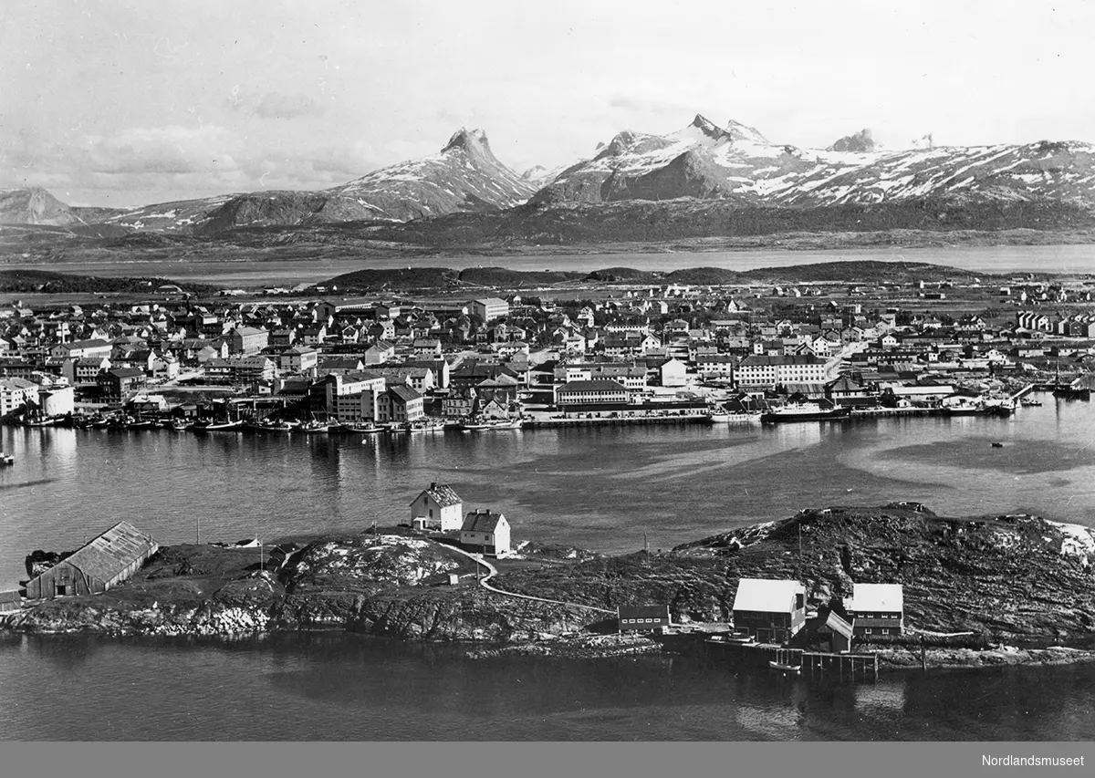 Nærbilde landskapet på Nyholmen sett fra Hjertøya med Bodø i bakgrunnen. Børvasstindene helt i bakgrunnen. I forgrunnen til venstre kull-lageret til Saltens Dampskipselskap. Bygningene i midten tilhører Fyrvesenet, til venstre en steinbygning, oppført 1875, det første fyret ved innseilinga til Bodø havn. Det andre er et trehus, oppført som bolig for Fyr- og Merkevesenets folk. Ved bryggene til venstre i forgrunnen er Merkevesenets lager.