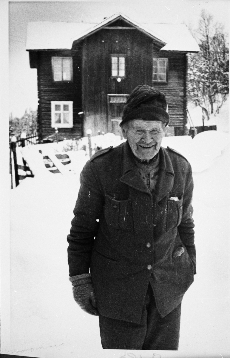 Portrett av en mann stående foran et hus. Mannen er iført lue og vinterklær.