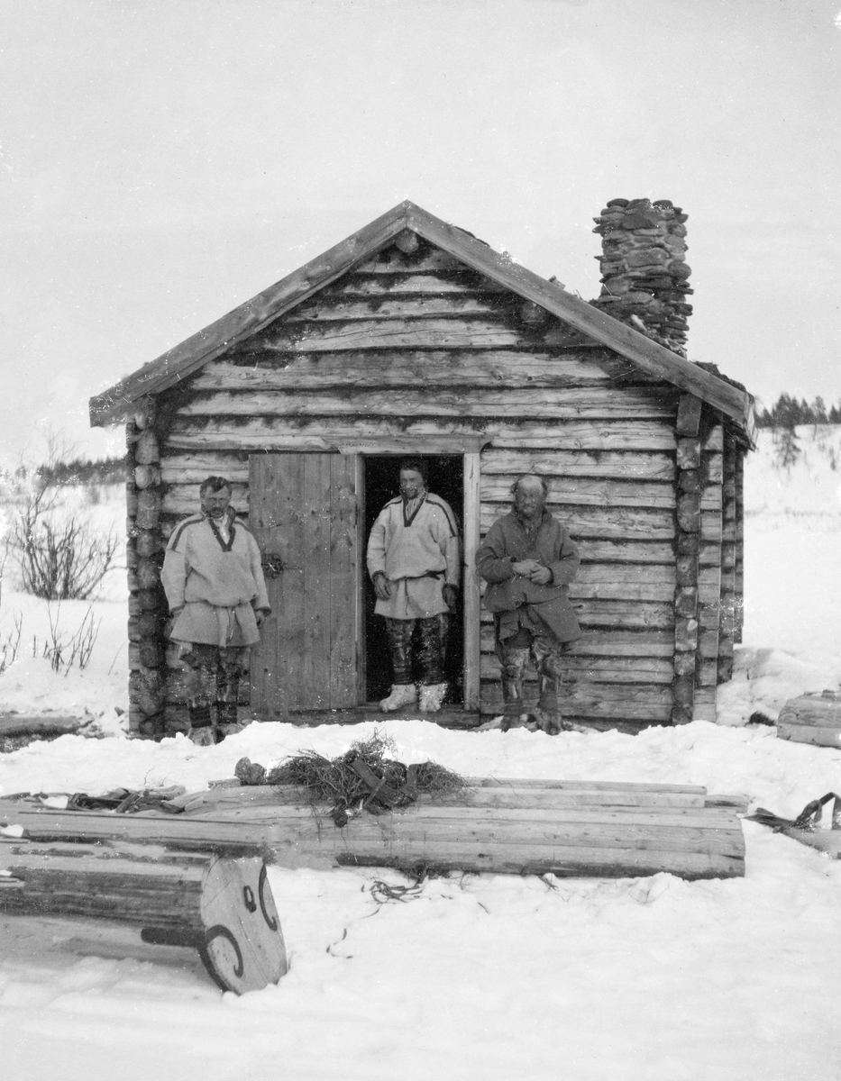 Tre samiske menn ved skogstua i Bassevnovde i Karasjokk.  De tre mennene står ved gavlveggen på bygningen, den midterste i døråpningen.  Stua er laftet og har gråsteinspipe.  I forgrrunnen ligger tre pulker med undersidene vendt i været.  Oppå to av dem ligger det en del sennegrasliknende fibrer. 