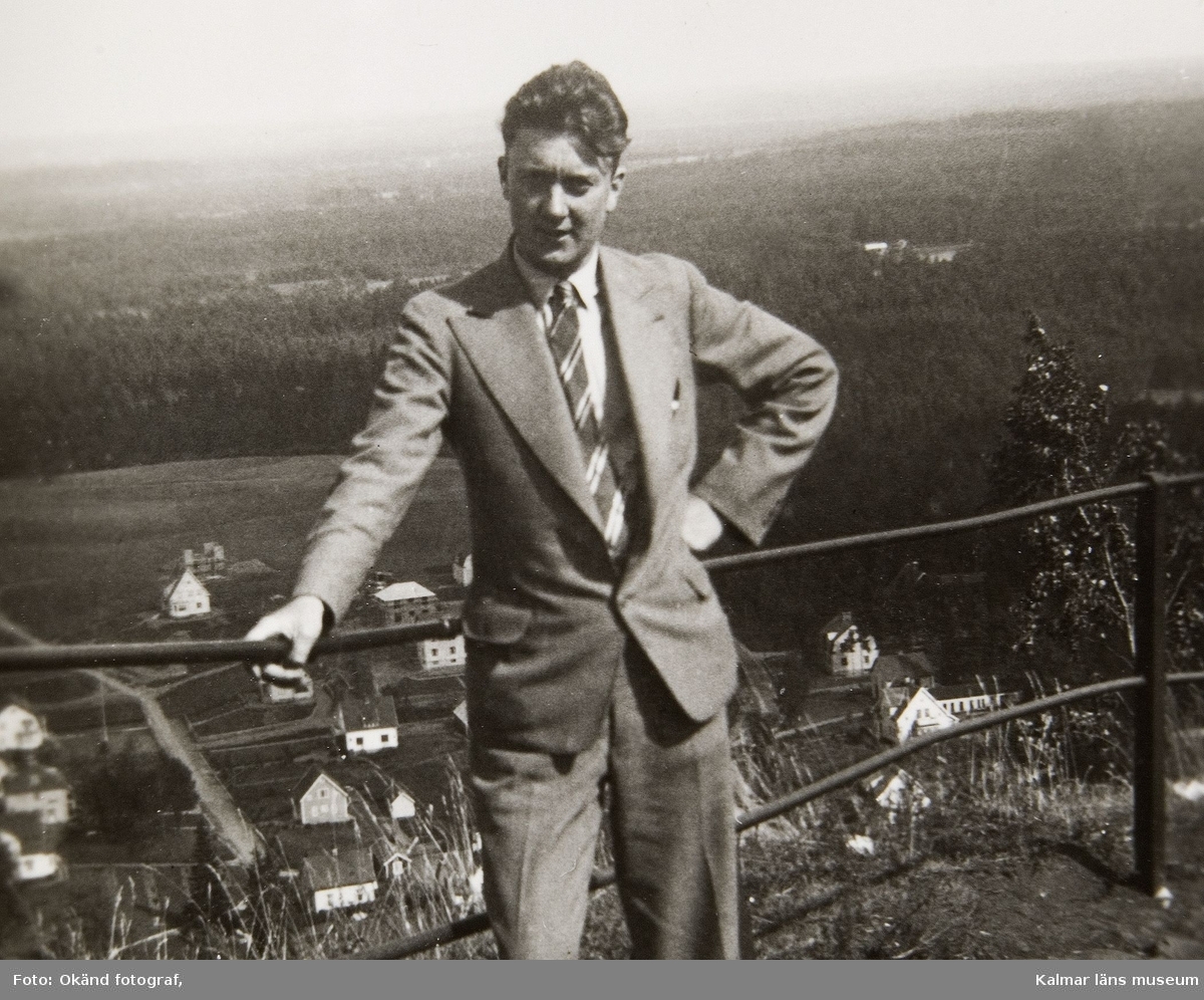 En man, troligtvis Valter Andersson, klädd i kostym, vit skjorta och randig slips står på en höjd vid ett räcke. Nedanför syns bebyggelse.