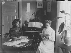 To kvinner sittende ved et bord i stue, en av kvinnene leser