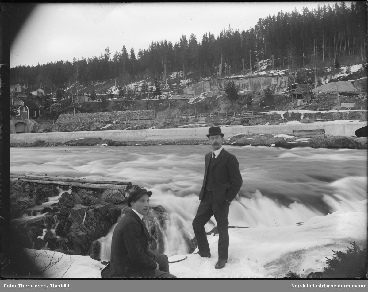To menn foran Sagafoss anlegget. Dammen delvis bygget i bakgrunnen. Boliger på oversiden av dammen.