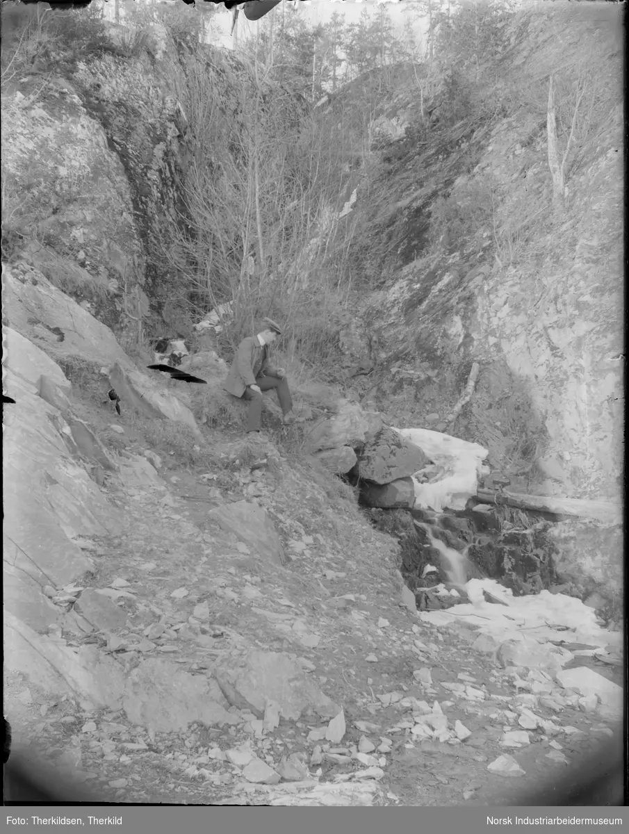 Mann sittende i skråning ved bekkefar, muligens ved Murukleivhola eller Hollagruvene.