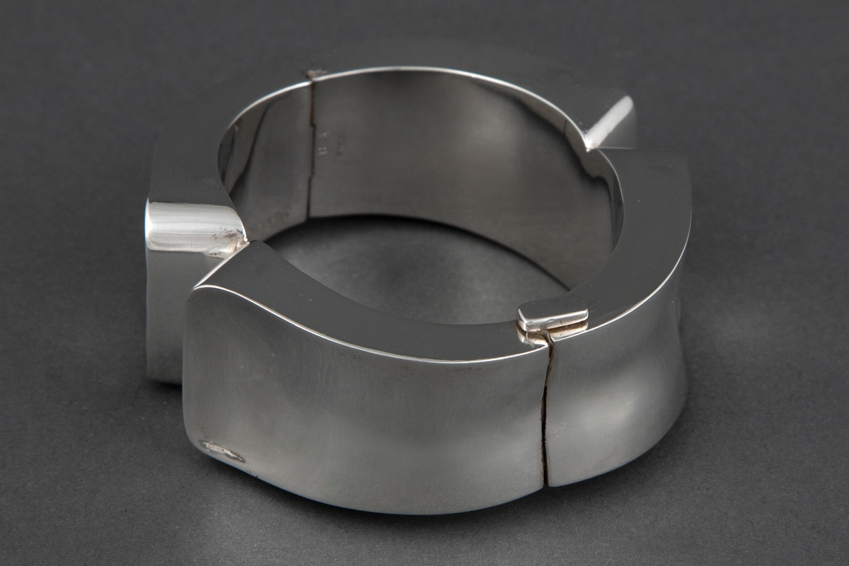 Kraftig armbånd med lukket form, bestående av to like deler som er forbundet med hengsel og lås. Yttersider med stor, konkav form i smidd sølv og innerside med glatt plate.