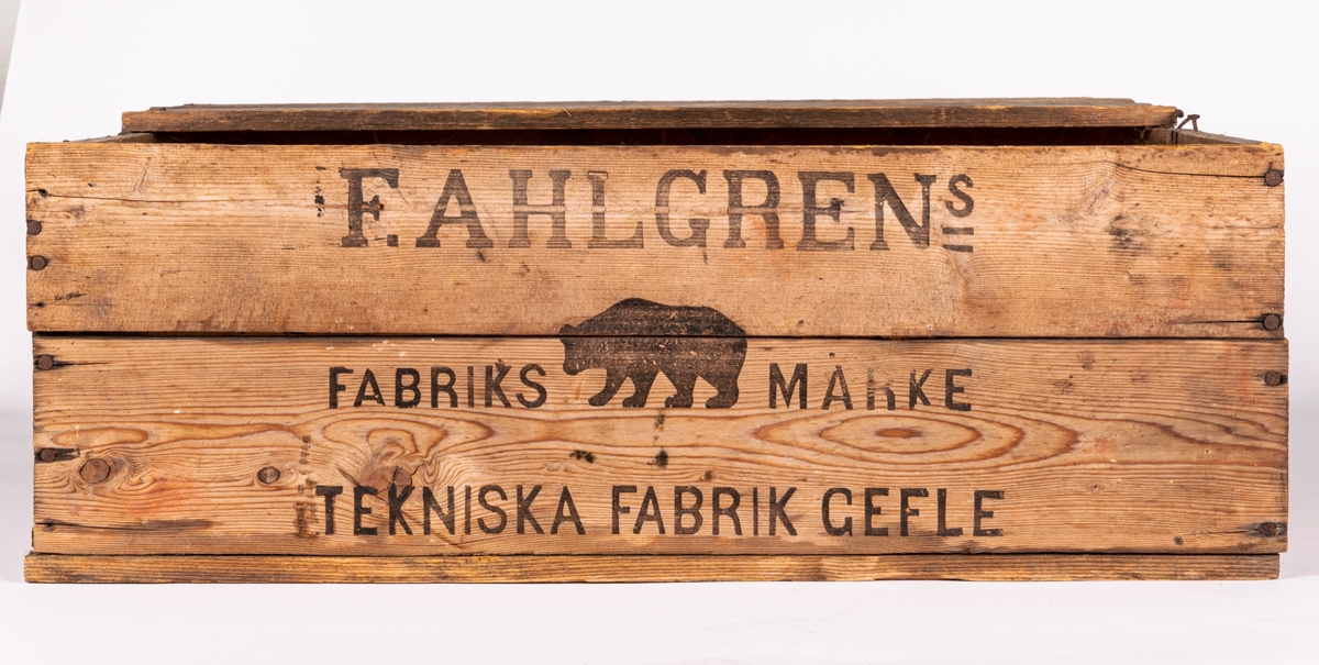 Trälåda, snickrad av ohyvlade brädlappar, spikad. Locket uppbrutet. Stämplar och påskrift på sidorna: "F. Ahlgrens Tekniska Fabrik".