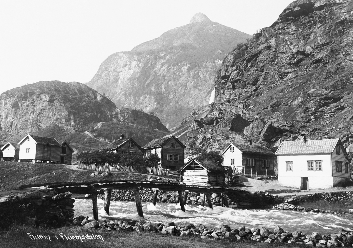 Bro over stri elv til bebyggelse i bygda Flåm innerst i Aurlandsfjorden med ruvende fjell i bakgrunnen, 1914.