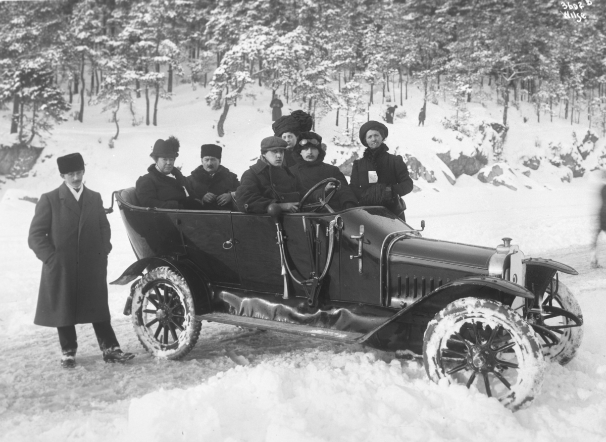Tre kvinner og fire menn i åpen bil i vinterlandskap. Ifølge protokoll tilhører bilen en pølsemaker Christiansen fra Christiania. Fotografert 1912.