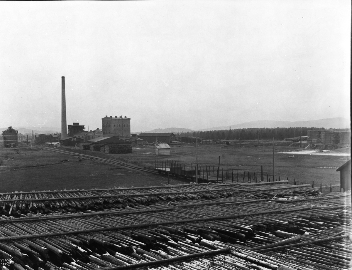 Masse tømmer ligger lagret foran fabrikkområde, Lillestrøm cellulosefabrikk. Fotografert 1923.