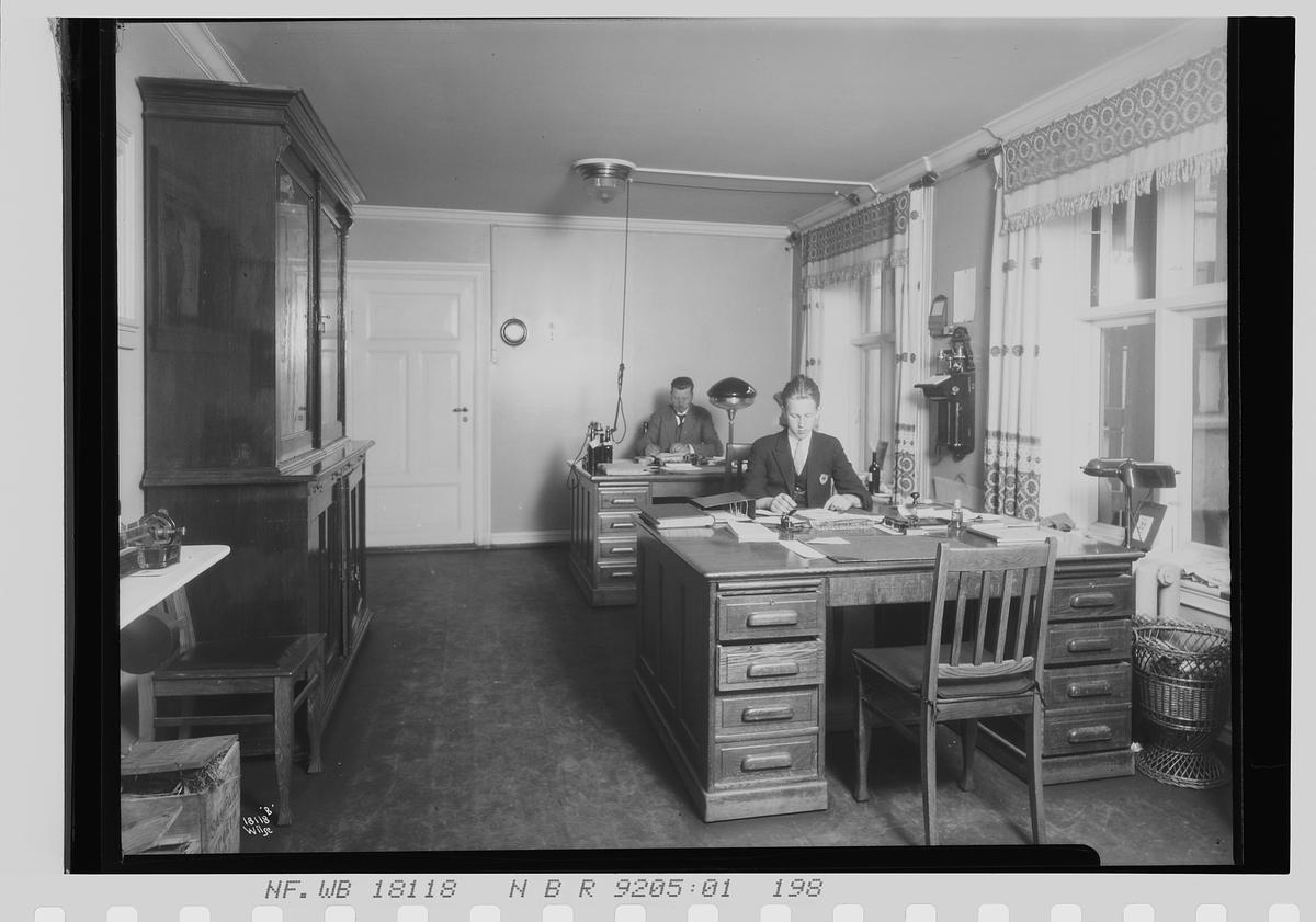 To menn sitter ved pultene på et kontor, Vinmonopolet. Fotografert 1924.