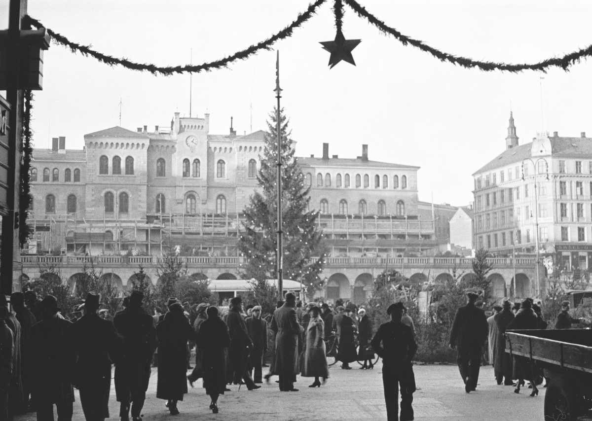 Youngstorget er julepyntet, det selges juletrær og tilstrømningen av byens beboere er stor.  I bakgrunnen troner Møllergata 19 og mot Youngstorget ser vi buegangene med kafeer og butikker av forskjellig slag.