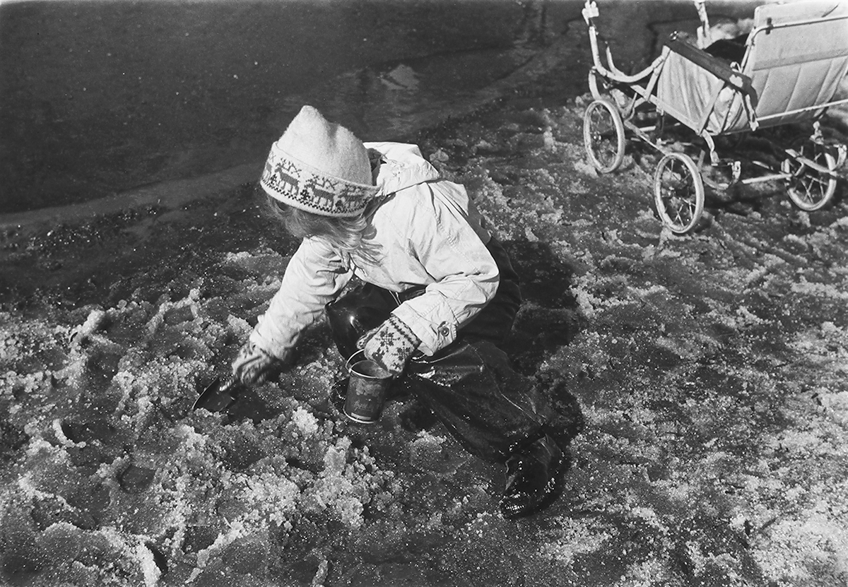 Pike med bøtte og spade i snøslaps. Barnevogn i bakgrunn. Fotografert 1940.
