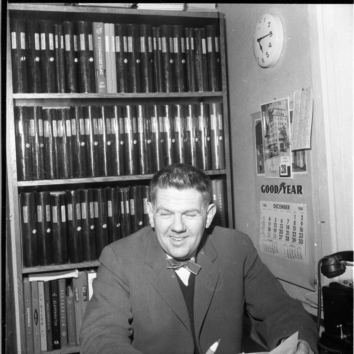 En man sitter på ett kontor. Bakom sig har han en bokhylla fylld med pärmar. Han ser glad ut.