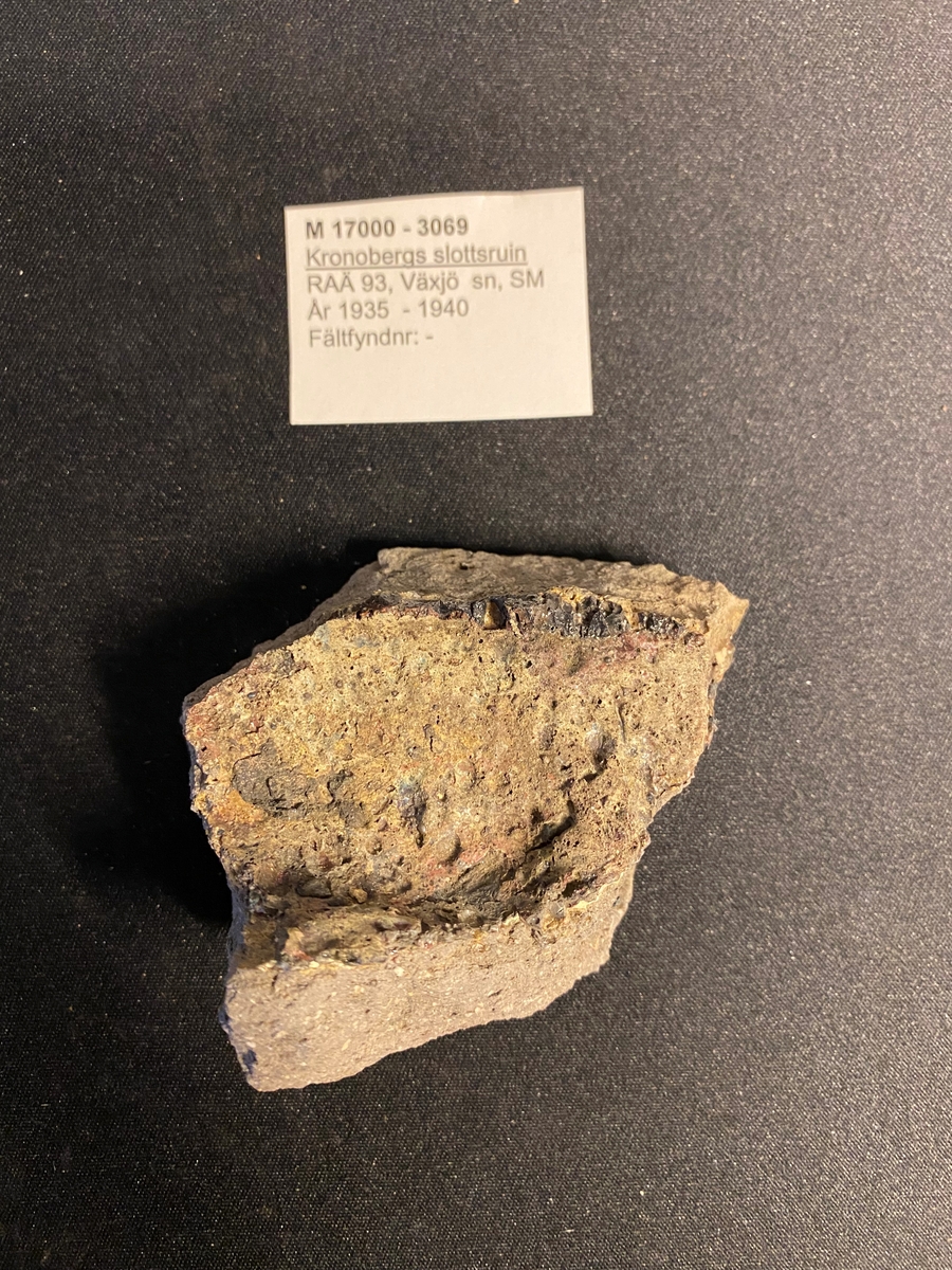 Fragment av botten till en degel. Sekundärt bränd med delvis cintrade röda och gröna ytor på den kvarvarande delen av buken. Relativt grovt gods. Liknar M 17000-3067.